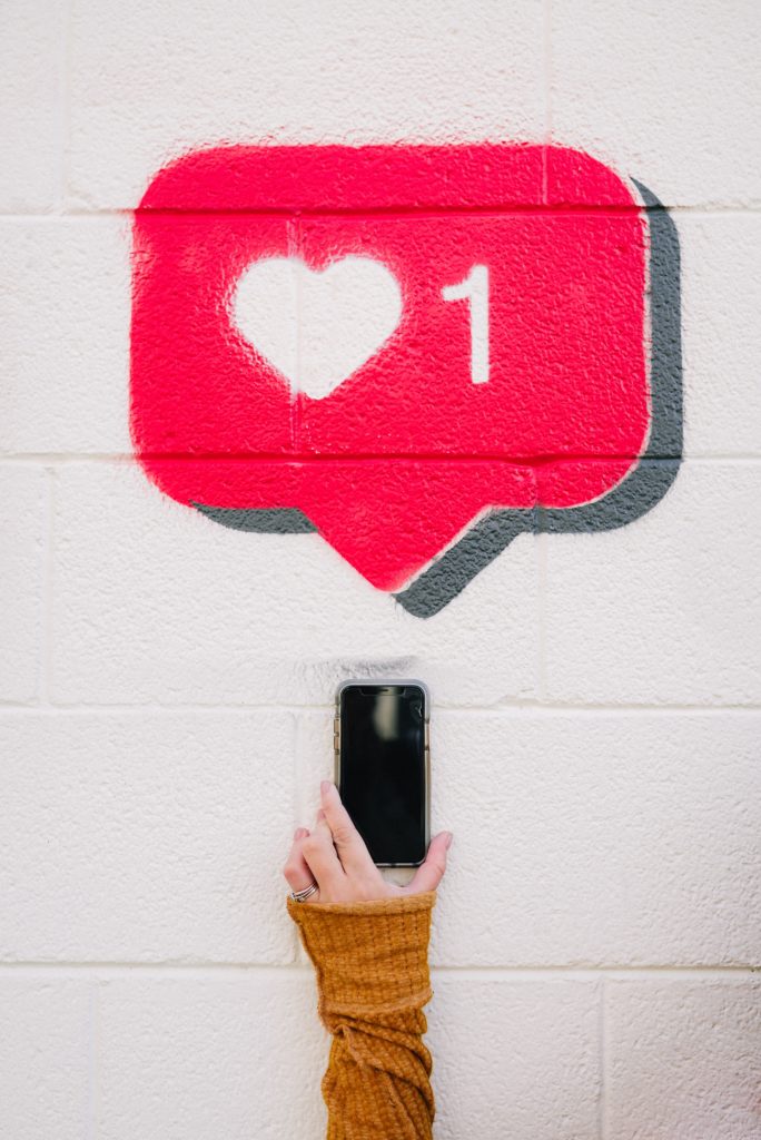 Title image for 10 Minute Marketing podcast episode "How To Write The Perfect Instagram Caption" depicting a hand holding up a smartphone under a red "1 Like" heart button painted on a white wall.