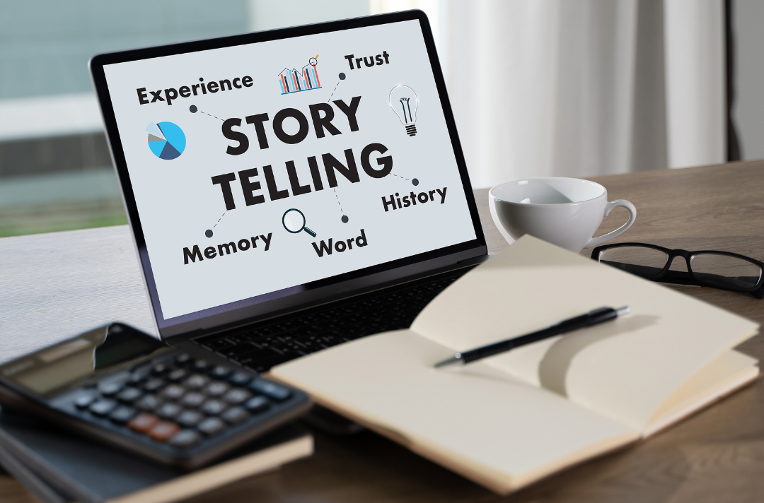 The Art of Storytelling Through Digital Marketing blog title card depicting a laptop with the words "Storytelling," "Experience" "Trust," "Memory," "Word," and "History" on its screen, sitting on desktop with an open notebook, a calculator, and a teacup.