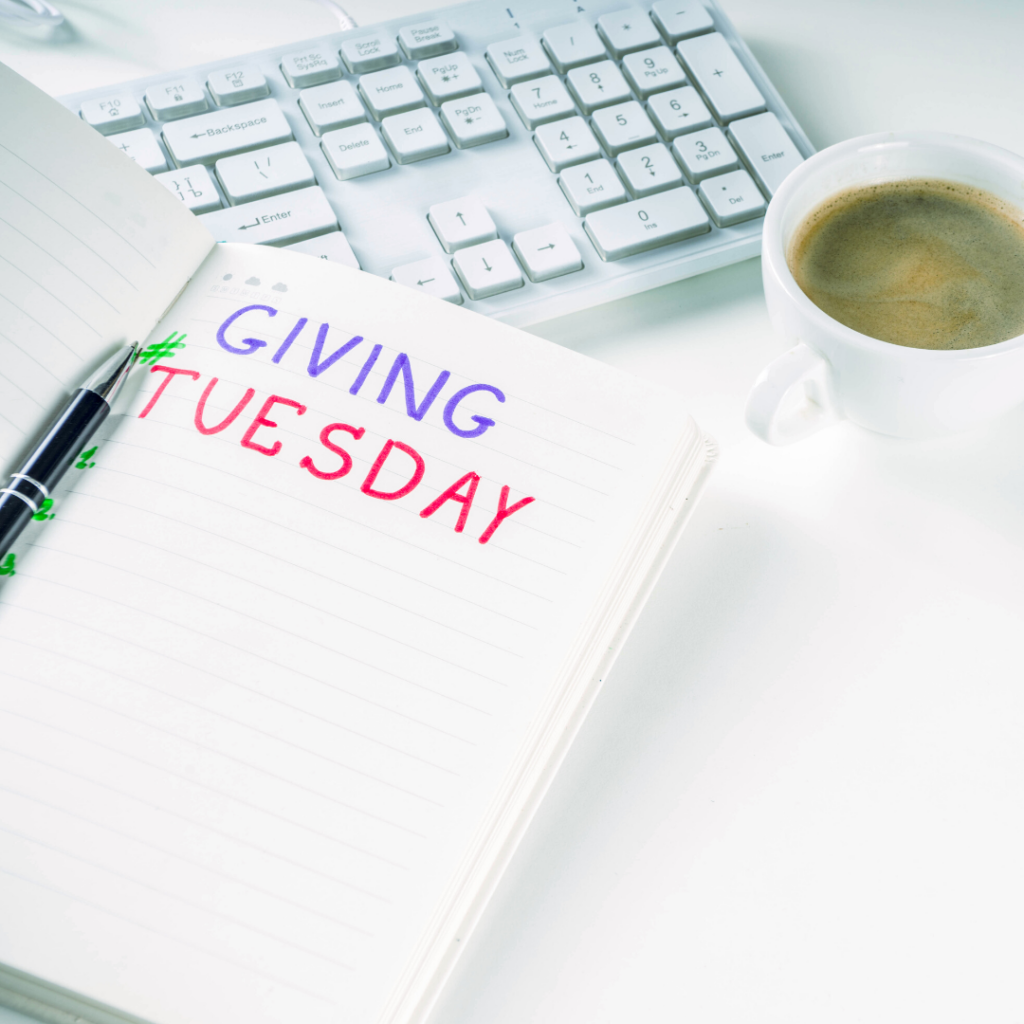 Black pen in the middle of an open notebook with "Giving Tuesday" written in blue and red ink, laying over a white keyboard on a white surface, with a white cup of coffee next to it.